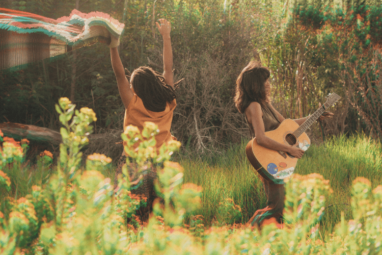 Two women enjoying nature together—one playing guitar, the other dancing—capturing the essence of joy, freedom, and true friendship