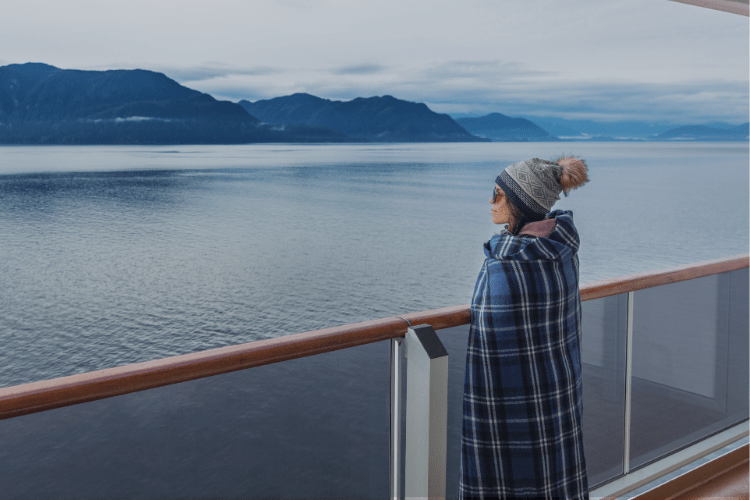 A woman wrapped in a blanket gazes over calm Alaskan waters, symbolizing solitude, reflection, and emotional connection