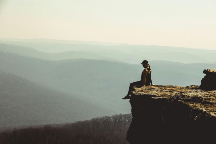 A woman sitting quietly on a cliff ledge, overlooking misty hills, reflecting calm, courage, and inner connection
