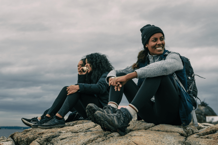 Three women sitting on a rocky ledge in hiking gear, laughing and resting after a climb, capturing adventure, trust, and friendship in motion