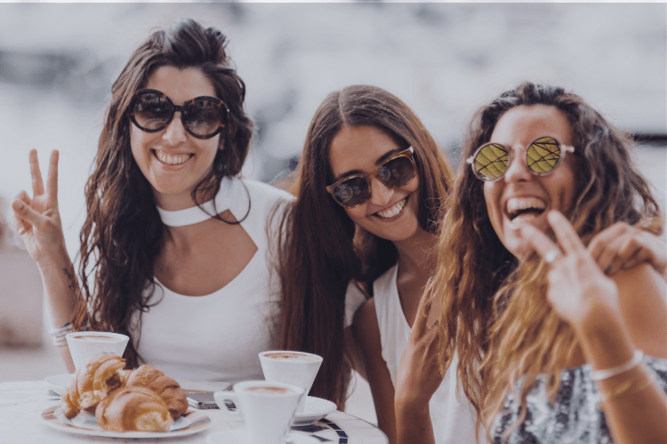 Three women laughing together at an outdoor café table, surrounded by coffee and croissants, radiating joy, friendship, and carefree connection