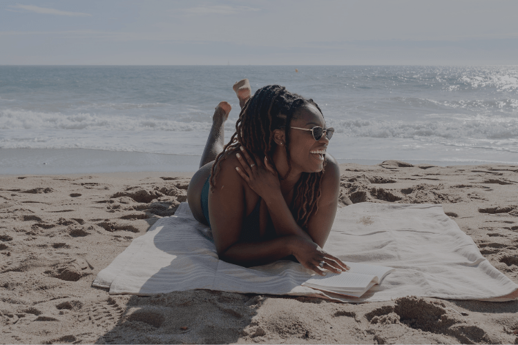 A woman lying on a beach towel, smiling brightly with a book beside her, enjoying the ocean breeze and a moment of carefree joy