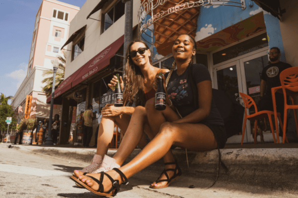 Two women sitting on a sunny sidewalk in Little Havana, holding drinks and laughing, surrounded by vibrant street life and colorful buildings